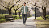 Homme portant une veste kimono vert sauge et un pantalon japonais moderne, marchant dans un parc fleuri au printemps sous les cerisiers