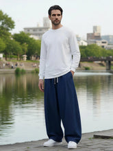 Pantalon japonais en jean pour homme porté debout en extérieur au bord d’une rivière, denim bleu foncé, coupe ample et fluide, taille à cordon ajustable, style minimaliste inspiré du Japon, silhouette élégante et naturell