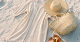 Kimono de plage écru, chapeau de paille et panier posés sur le sable clair