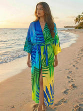Kimono long bohème motif palmier bleu et vert porté par une femme sur une plage tropicale au coucher du soleil