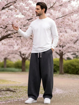 Pantalon japonais en jean pour homme porté debout en extérieur sous des cerisiers en fleurs, denim gris foncé, coupe ample et fluide, taille à cordon ajustable, style minimaliste inspiré du Japon, ambiance naturelle et poétique.