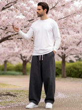 Pantalon japonais en jean pour homme porté debout en extérieur sous des cerisiers en fleurs, denim gris foncé, coupe ample et fluide, taille à cordon ajustable, style minimaliste inspiré du Japon, ambiance naturelle et poétique.