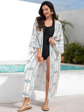 Kimono de plage long femme blanc à motifs géométriques bleus, porté ouvert sur un maillot noir, style estival élégant au bord d’une piscine.